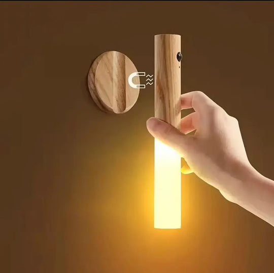 A hand holds a glowing cylindrical wooden wall light against a warm brown wall, with a round wooden mounting plate shown beside it and a small magnet graphic indicating magnetic interaction.